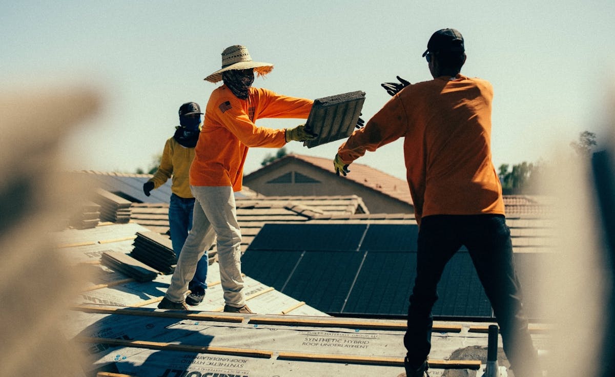 Roofing crew passing tiles while working on a residential roof under sunny weather, wearing protective gear and safety clothing.