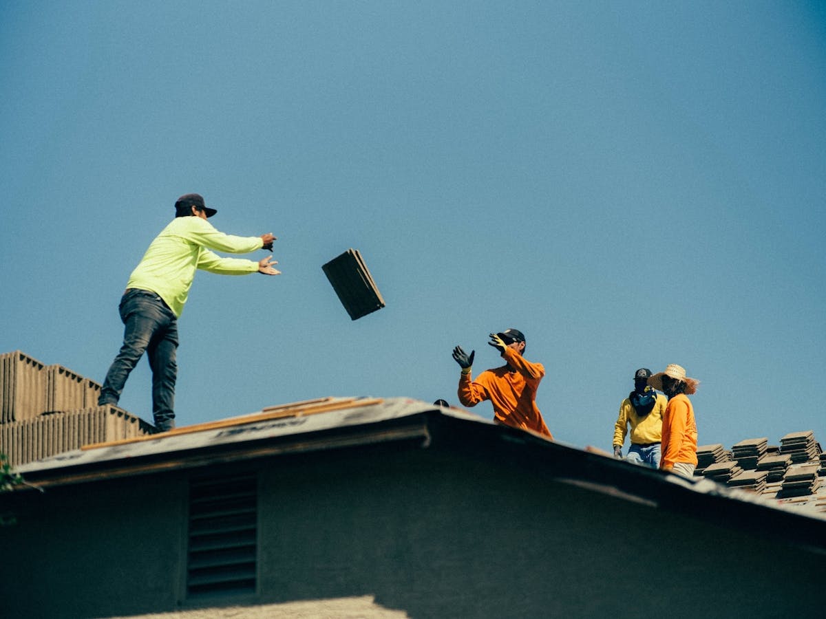 construction workers on a rooftop pass roofing tiles by hand with one worker mid-throw and others preparing to catch or stack the materials