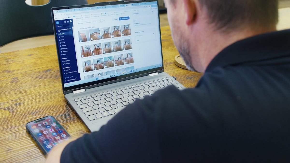Contractor reviewing construction software dashboard with job site photos on a laptop in an office conference room.